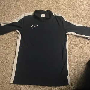 Marinblå Nike half zip tröja - Sportig marinblå långärmad tröja från Nike med vit logga på bröstet och vita paneler längs ärmarna. Tröjan har en half zip-dragkedja vid halsen och är gjord i mjukt syntetmaterial som passar perfekt för träning eller chill.