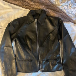 Svart skinnjacka från Maryland Outwear - Snygg svart skinnjacka från Maryland Outwear i storlek S. Jackan har raka linjer, klassisk krage och dragkedja framtill. Två fickor framtill och stilren design som passar perfekt till streetwear eller en edgy look.