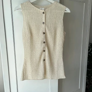 Beige ärmlös blus från H&M - Säljer en beige blus från H&M i storlek S. Toppen är ärmlös med rund hals och har ett vågigt, strukturerat tyg. Den är helt oanvänd med lappen kvar!