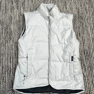 Vit dunväst från Tenson - Vit dunväst från Tenson med hög krage och dragkedja framtill. Västen har quiltad design och är tillverkad i vattenavvisande och andasvänligt material. Snygga broderade detaljer och logga på sidan. Perfekt för lager-på-lager under kyliga dagar.