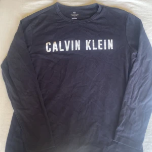 Mörkblå långärmad tröja Calvin Klein - Snygg mörkblå långärmad tröja från Calvin Klein Performance med vit logotyp tryckt över bröstet. Tröjan har en klassisk rund hals och är gjord i mjuk bomull, perfekt för chill dagar eller när du vill ha en clean look.