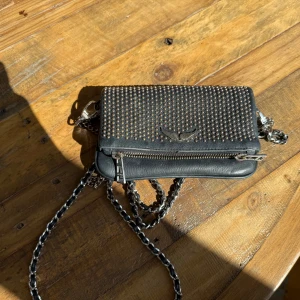 Svart axelväska med nitar från ZADIG & VOLTAIRE - Cool svart axelväska i skinn från ZADIG & VOLTAIRE med silverfärgade kedjor och massor av små nitar på framsidan. Väskan har en dragkedja framtill och ett snyggt metallmärke. Perfekt storlek för mobilen och det viktigaste. ÄKTA köptes för 3999 säljer nu för 2000 kr men priset kan diskuteras 