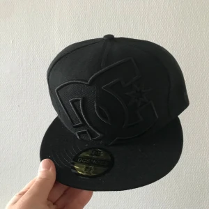 Svart DC Shoes keps 7 3/8 - Säljer en svart keps från DC Shoes med stort broderat DC-logo framtill. Klassisk flat brim och sticker på skärmen. Kepsen är i helsvart och har en clean streetstyle-look, perfekt för dig som gillar skate eller urban stil.