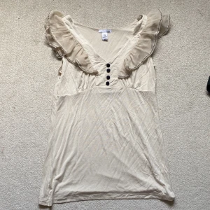 Beige volangblus från H&M - Supersöt beige blus från H&M med volangdetaljer längs axlarna och v-ringning. Dekorerad med fyra svarta knappar framtill och sydd i ett mjukt, lätt material. Perfekt för dig som gillar romantiska och feminina detaljer.