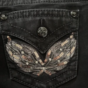 Svarta Miss Me jeans med paljettdetaljer - Säljer ett par svarta jeans från Miss Me med snygga broderade bakfickor dekorerade med paljetter och stenar. Jeansen har låg midja, bootcut och coola knappar med mönster. Midjemått:37cm innerben:75cm