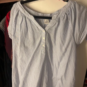 Randig blus från Isolde i bomull - Säljer en ljusblå och vit randig blus från Isolde i storlek S. Blusen har korta ärmar, v-ringning och knappar framtill. Tillverkad i mjuk bomull, perfekt för en chill och fräsch look.