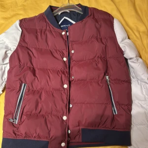 Bordeaux pufferjacka från GANT - Snygg pufferjacka från GANT i bordeaux med kontrasterande vita ärmar. Jackan har quiltad design, silverfärgade dragkedjor på fickorna och tryckknappar framtill. Ribbad mudd vid krage och nederkant ger en sportig vibe. Perfekt för kyliga dagar.