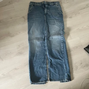 Jeanskjol med slits från NA-KD - Snygg lång jeanskjol från NA-KD med en stilren design och en slits framtill. Kjolen har en klassisk blå denimfärg och är perfekt för en avslappnad look. Den har en traditionell femficksdesign och bälteshällor i midjan.