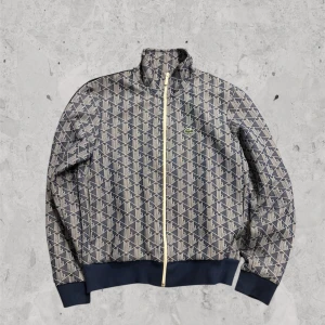 Lacoste monogram  - Säljer en snygg mönstrad jacka från Lacoste med dragkedja och långärmade ärmar. Jackan har ett geometriskt mönster i blått och beige och är tillverkad i en blandning av polyester och bomull. Perfekt för en stilren look. Köpt på johnells för ca 4 månader sen men har inte kommit till användning har kvar påse men inte kvitto, vid snabb affär kan jag gå ner lite i pris. Inga defekter (nytt skick) og pris 2500