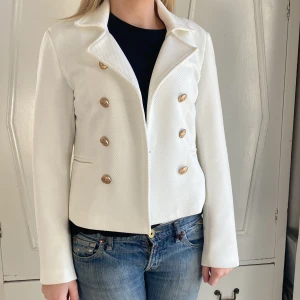 Vit kavaj med guldknappar från Isay - Fin helfordrad vit kavaj med stiliga guldknappar, åtta fram och tre på varje ärm. Kavajen ser dubbelknäppt ut men knäpps med en hyska och hake mitt fram. Använd vid ett tillfälle. Spår av brun utan sol kan skymtas i nacken på insidan av kragen. Längd 56. Storlek 38. 