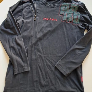 Svart långärmad tröja från Prada - Säljer en stilren svart långärmad tröja från Prada med röd logotyp på bröstet och en unik dragkedjedetalj vid axeln. Tröjan har även ett intressant mönster i grönt och rött på axeln. Perfekt för dig som vill ha något exklusivt och trendigt i garderoben.