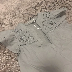 Grön blus med broderad detalj - Säljer en söt grön blus med vackra broderade detaljer vid halsen. Blusen har korta ärmar och en avslappnad passform. Perfekt för en stilren och bekväm look.