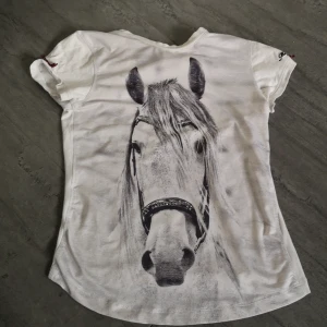 Vit t-shirt med hästmotiv från Mountain Horse - Säljer en vit t-shirt från Mountain Horse med ett stort hästmotiv på framsidan. T-shirten har korta ärmar och två små rosetter på ärmarna. Perfekt för hästälskare! 🐴 I bra skick, inga hål och bara andvände få gånger. 