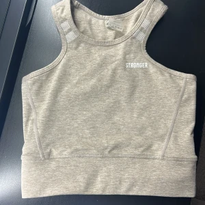 Beige linnetopp från Stronger - Snygg beige linnetopp från Stronger med vita detaljer och text. Perfekt för träning med sin bekväma passform och stilrena design. Toppen har en hög halsringning och ärmlös design. Nästan aldrig använd 