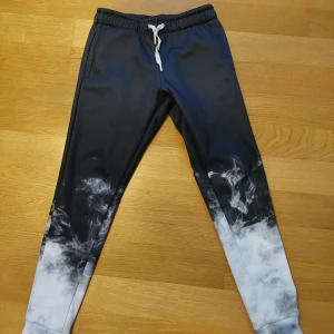 Svarta mjukisbyxor med rökdesign från Mr. Gugu & Miss Go - Coola svarta mjukisbyxor med unik rökdesign i vitt och grått. Byxorna har en elastisk midja och är perfekta för en avslappnad stil. De är tillverkade i ett mjukt material som ger en bekväm passform.