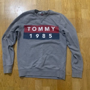 Grå sweatshirt från Tommy Jeans - Säljer en grå sweatshirt från Tommy Jeans med tryck 'TOMMY 1985' i rött och blått på framsidan. Tröjan har långa ärmar och en rund halsringning med ribbade muddar vid ärmslut och nederkant.