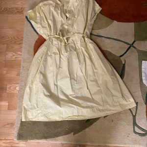 Ljusgul klänning från Uniqlo - Säljer en ljusgul klänning från Uniqlo med knytband i midjan. Klänningen har korta ärmar och en avslappnad passform. Finns några små fläckar nere i kanten på framsidan men är inte något som jag tycker syns tydligt när man använder den 