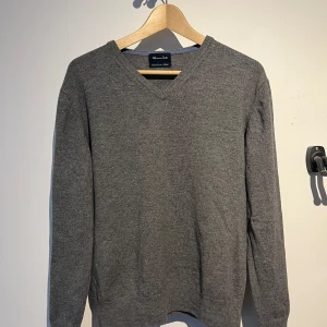 Grå tröja från Massimo Dutti - Säljer en stilren grå V-neck tröja från Massimo Dutti. Tröjan har en v-ringad design och är tillverkad i Cashmere. Den är i bra skick förutom ett mindre hål på högra armen, därav priset. Tröjan passar ca. 175. Hör av er vid frågor!