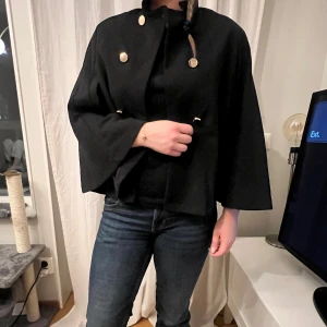 Svart kappa med knappar - Elegant svart kappa med stora dekorativa knappar. Den har en lös passform och trekvartsärmar, vilket ger en stilren och modern look. Perfekt för både höst och vår. Säljer för den inte kommer till användning.