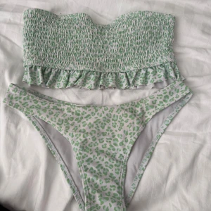 Grön leopardmönstrad bikini - Trendig bikini med grönt leopardmönster. Överdelen är axelbandslös med smockdetaljer och volangkant. Underdelen har en klassisk trosa med samma mönster. Perfekt för sommarens stranddagar!