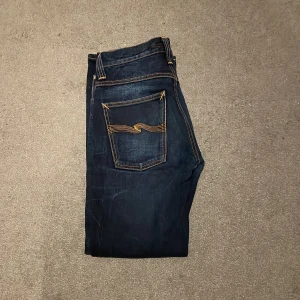 Mörkblå jeans från Nudie Jeans - Säljer ett par mörkblå jeans från Nudie Jeans med klassiska orangea sömmar. Jeansen har en normal passform och är tillverkade i slitstarkt denim. Perfekta för en stilren look.
