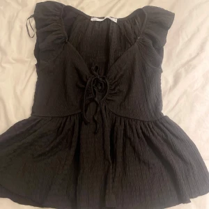 Svart topp med knyt från 157 - Snygg svart topp från 157 med knytning framtill och volangdetaljer vid ärmarna. Toppen har en lätt rynkad textur och en luftig passform som ger en avslappnad look. Perfekt för en stilren outfit.