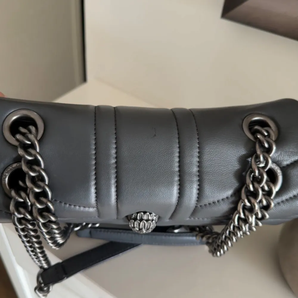 Snygg grå väska från Kurt Geiger i skinn med kedjedetaljer. Väskan har en elegant design med ett dekorativt emblem på framsidan och en praktisk insida. Väskan går borde att bära under axeln eller som en axelremsväska💛💛. Laukut & Käsilaukut.