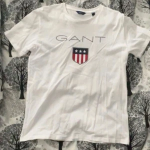 Vit t-shirt från GANT - Säljer en vit t-shirt från GANT med ett tryck av deras logga och ett sköldemblem i rött, vitt och blått. Perfekt för en stilren och avslappnad look. Passar ungdomar i åldern 13-14 år.