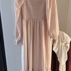 Ljusrosa långklänning med smockdetaljer - Charmig ljusrosa långklänning med smockdetaljer upptill och långa ärmar. Klänningen har en volangkant vid fållen och elastiska ärmslut för en bekväm passform. Perfekt för en romantisk look.