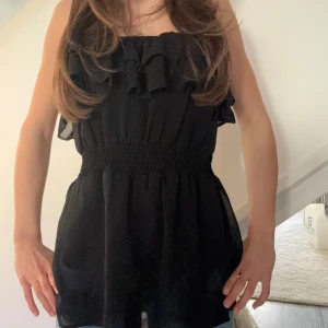 Svart axelbandslös klänning - Snygg svart axelbandslös topp  med volangdetaljer upptill. Den har en elastisk midja som ger en smickrande passform. Perfekt för en stilren look. 