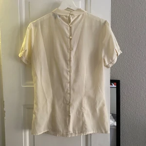  Blus med knappar - Säljer en stilren cremefärgad / vit  blus med korta ärmar och knappar framtill. Blusen har en elegant design med en knytbar detalj baktill. Perfekt för en chic look!