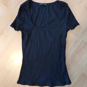 Svart ribbad topp från Zara - Säljer en stilren svart ribbad topp från Zara. Toppen har en djup rundad halsringning och korta ärmar. Perfekt för lager-på-lager eller att bära som den är. Säljer för att jag råkade köpa den i fel storlek, storlek S