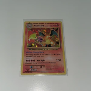 Charizard Pokémon-kort - Säljer ett Charizard Pokémon-kort med 150 HP och förmågan Energy Burn. Kortet har en Fire Spin-attack som gör 200 skada. Det är ett holografiskt kort med en färgglad bakgrund och är skyddat i en plastficka.