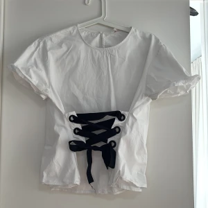 Vit blus med svart snörning - Säljer en stilren vit blus med korta ärmar och en snygg svart snörning framtill. Perfekt för att ge en touch av elegans till din outfit. Blusen har en rund halsringning och är tillverkad i ett lätt och bekvämt material.