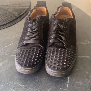 Svarta Louboutin - Säljer ett par svarta sneakers med nitar på tån och ovansidan. Skorna har snörning och en röd sula. Perfekta för dig som vill ha en edgy stil.