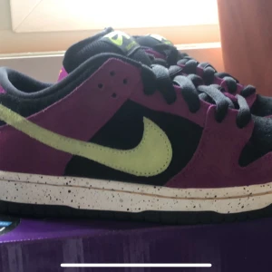 Nike SB Dunk Low Pro red plum - Snygga Nike SB Dunk Low Pro i en cool färgkombination av svart, lila och lime. Skorna har en kontrasterande sula med stänkdetaljer och klassisk snörning. Perfekta för dig som vill sticka ut med stil. En fin sko från restocks i perfekt skick. Skon är lite använd och är i storlek 38,5. Låda och extra skosnören finns. Beviset på att dem är äkta är från taggen från restocks, lådan, och skon i sig. Bara att fråga om fler bilder. Priset kan diskuteras!