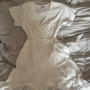 Vit studentklänning - Säljer en elegant vit omlottklänning med korta ärmar och volangdetaljer. Klänningen har en v-ringad design och knytband i midjan. Jättefin, använd en gång, som ny. Går ovanför knäna, sitter ännu finare på. Passar xs-s då jag brukar använda S annars. Köpt för 500. Använd en gång på studenten