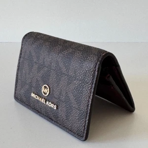 Brun plånbok från Michael Kors - Snygg brun plånbok från Michael Kors med elegant logotyp i guld på framsidan. Plånboken har en praktisk knappstängning och flera fack för kort och ID. Insidan är i en stilren vinröd färg som ger en lyxig känsla.