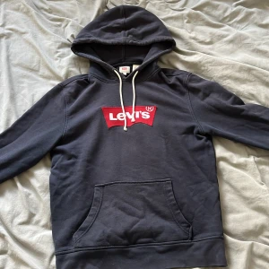 Mörkblå hoodie från Levi's - Säljer en mörkblå hoodie från Levi's med det klassiska röda logotyptrycket på bröstet. Tröjan har en justerbar huva med vita snören och en praktisk magficka framtill. Perfekt för en avslappnad stil.