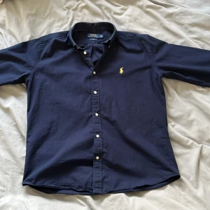 Mörkblå skjorta från Polo Ralph Lauren - Snygg mörkblå skjorta från Polo Ralph Lauren med klassisk krage och knappar framtill. Skjortan har en broderad gul logga på bröstet och är i slim fit. Perfekt för en stilren look.