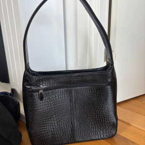 Svart axelväska med krokodilmönster - Snygg svart axelväska med krokodilmönster och dragkedja framtill. Perfekt för att ge en stilren touch till din outfit. Väskan har en bekväm axelrem och gott om plats för dina nödvändigheter.