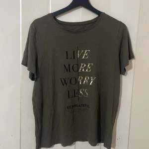 Olivgrön t-shirt med text - Säljer en olivgrön t-shirt med texten 'Live More Worry Less' i glittrande bokstäver. T-shirten har en rund hals och korta ärmar. Perfekt för en avslappnad stil.
