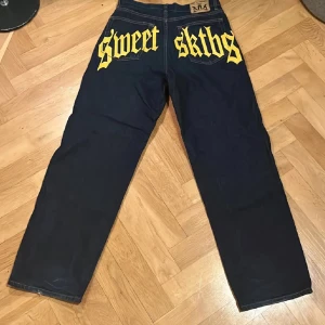 Svarta jeansbyxor från Sweet SKTBS - Säljer ett par svarta jeansbyxor från Sweet SKTBS med fett gult tryck på baksidan. Passar ungefär 170 och uppåt. Byxorna har en normal passform och är perfekta för en avslappnad stil. De har klassisk femficksdesign och knappgylf.