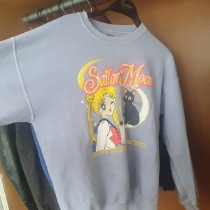 Grå Sailor Moon sweatshirt - Säljer en grå sweatshirt med ett färgglatt Sailor Moon-tryck på framsidan. Tröjan har en rund halsringning och långa ärmar. Perfekt för fans av serien!