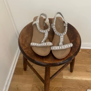 Silvriga sandaler med pärldetaljer storlek 39 - Snygga silvriga sandaler från Etnas med glittrande pärldetaljer. De har en bekväm kilklack och elastiska remmar för enkel passform. Perfekta för sommarens alla tillfällen.