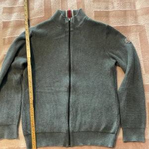 Grå stickad tröja från Moncler - Säljer en stilren grå stickad tröja från Moncler med dragkedja och ribbad krage. Tröjan har långa ärmar och ett Moncler-märke på ärmen. Perfekt för kyligare dagar.