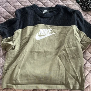 Olivgrön och svart t-shirt från Nike - Säljer en olivgrön och svart t-shirt från Nike med korta ärmar och ett stort vitt Nike-logotyptryck på framsidan. Den har en cool mesh-detalj på axlarna och är perfekt för en sportig look. Passar bra till både träning och vardag.