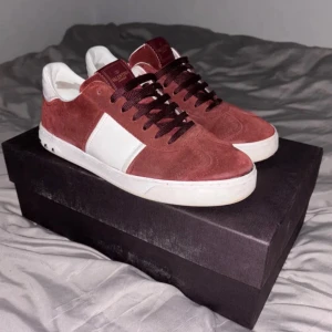 Röda mocka sneakers från Valentino - Snygga röda mocka sneakers från Valentino med vita detaljer och klassisk snörning. Skorna har en stilren design med märkets logga på plösen och nitar på hälen för en extra touch. Perfekta för dig som vill ha en stilren look med en twist.
