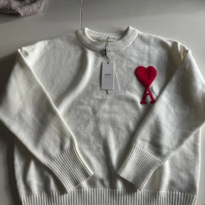 Vit tröja från Ami Alexandre Mattiussi - Säljer en stilren vit tröja från Ami Alexandre Mattiussi med ett rött hjärta och bokstaven A broderat på bröstet. Tröjan har en rund halsringning och ribbade muddar vid ärmslut och nederkant. Perfekt för en trendig look! 🖤