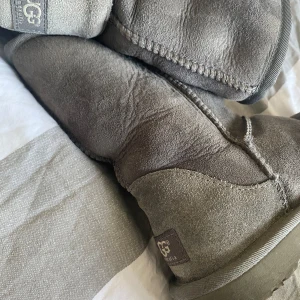Grå mocka boots från UGG - Säljer ett par klassiska grå boots från UGG i mjuk mocka. De har en rund tå och är perfekta för kyligare dagar. UGG-loggan syns tydligt på baksidan av stövlarna. Sulan är robust och ger bra grepp.